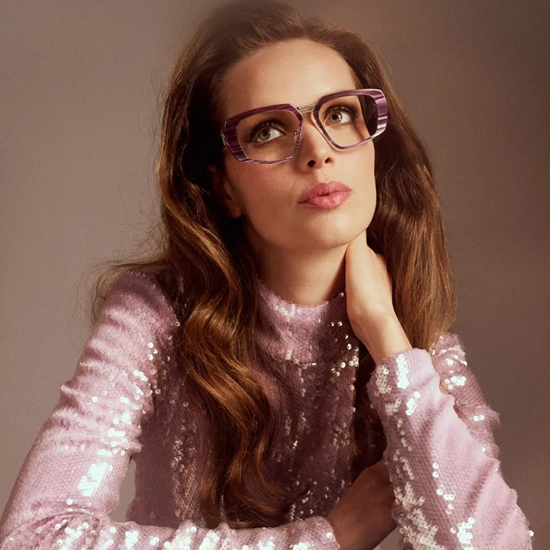 A woman in a glittery pink top is wearing the purple model Cassiopeja from Studio Eyewear